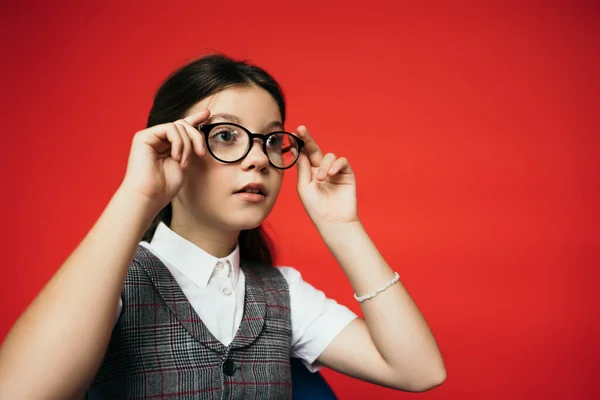Girl in plaid vest looking away and adjusting eyeglasses isolated on red — Stock Photo