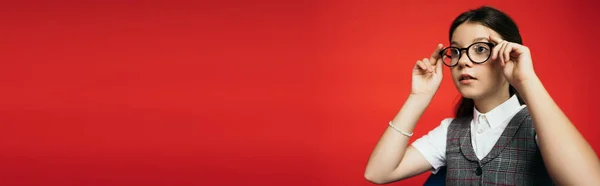 Brunette girl adjusting stylish eyeglasses and looking away isolated on red, banner — Stock Photo
