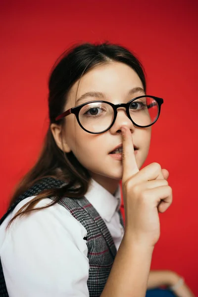 Portrait of girl in eyeglasses looking at camera and showing hush gesture isolated on red — Stock Photo