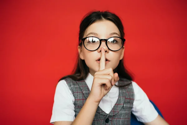 Portrait of preteen girl in eyeglasses looking at camera and showing shh sign isolated on red — Stock Photo