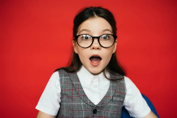 Astonished girl in eyeglasses and plaid vest looking at camera isolated on red — Stock Photo