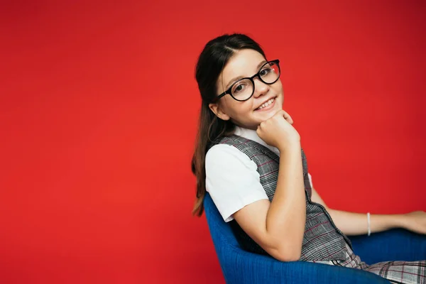 Menina satisfeito na moda óculos sentados em poltrona com a mão perto do queixo isolado no vermelho — Fotografia de Stock