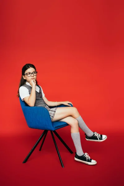 Longitud completa de niña reflexiva en zapatos de goma y anteojos sentados en sillón y mirando hacia otro lado sobre fondo rojo - foto de stock