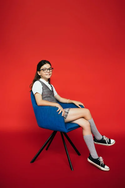 Полная длина улыбающейся девушки в очках и ботинках, сидящей в синем кресле на красном фоне — стоковое фото