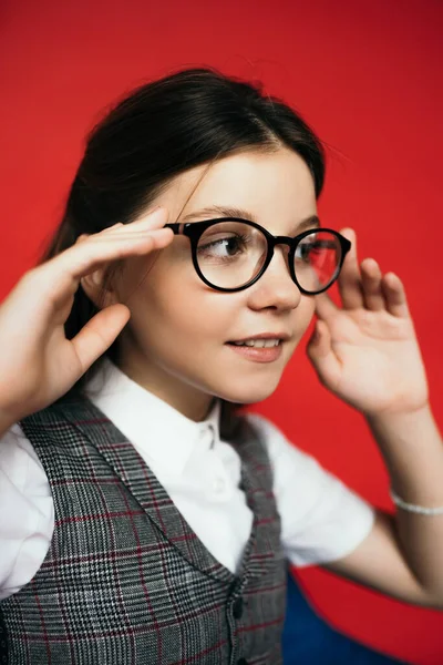 Preteen girl adjusting eyeglasses and smiling while looking away isolated on red — Stock Photo