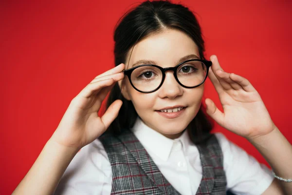 Lächelndes brünettes Mädchen, das eine Brille berührt und isoliert auf rot in die Kamera schaut — Stockfoto