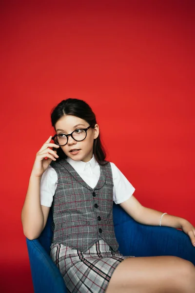 Fille en gilet à carreaux ajustant les lunettes à la mode tout en étant assis dans le fauteuil et regardant loin isolé sur rouge — Photo de stock