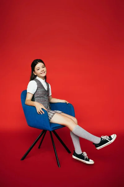Volle Länge des fröhlichen Mädchens in Kniestrümpfen und Gummischuhen sitzt im blauen Sessel auf rotem Hintergrund — Stockfoto
