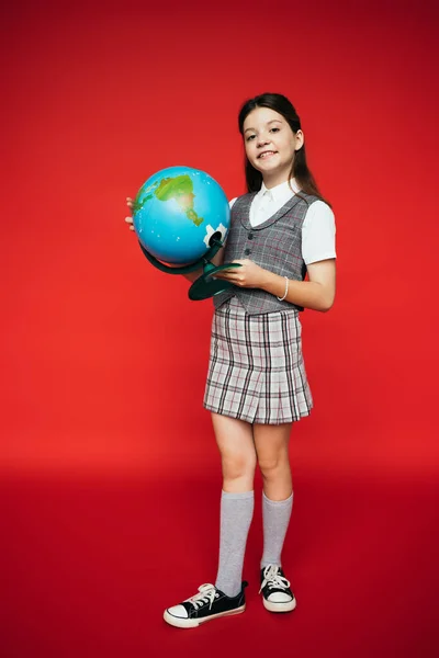 Повна довжина щасливої дівчини в плетеній спідниці і камерах позує з глобусом на червоному тлі — стокове фото