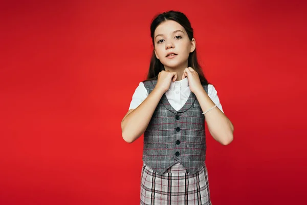 Preteen girl in checkered vest and skirt adjusting collar of white blouse isolated on red, banner — Stock Photo