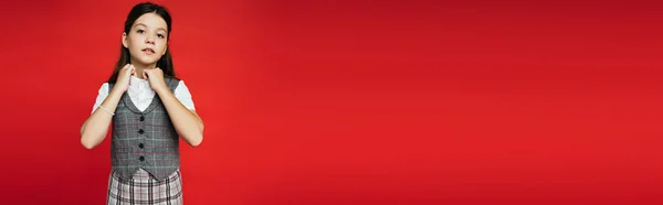 Брюнетка-подросток в клетчатом жилете регулируя воротник блузки и глядя на камеру изолированы на красный, баннер — стоковое фото