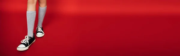 Abgeschnittene Ansicht von Mädchen in grauen Kniestrümpfen und trendigen schwarz-weißen Gummischuhen auf rotem Hintergrund, Banner — Stockfoto