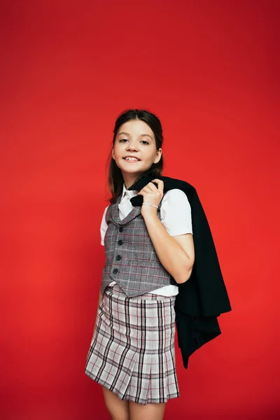 Brunette girl in checkered clothes holding black jacket and smiling at camera isolated on red — Stock Photo