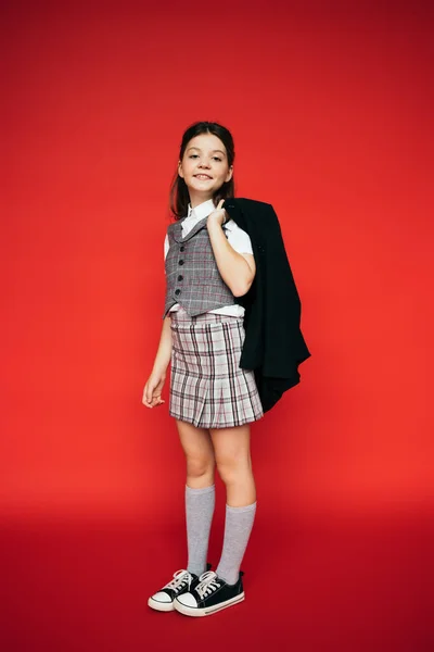 Volle Länge des lächelnden Mädchens in kariertem Rock und Gummischuhen mit schwarzer Jacke auf rotem Hintergrund — Stockfoto