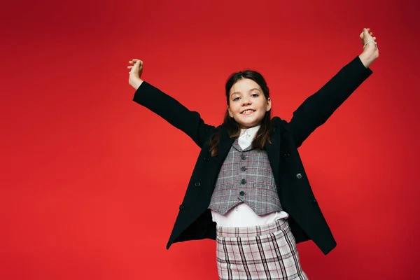 Cheerful girl in plaid vest and black blazer standing with outstretched hands isolated on red — Stock Photo