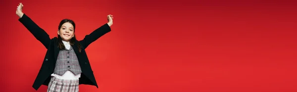 Fröhliches Mädchen in karierter Weste und schwarzem Blazer posiert mit ausgestreckten Händen auf rotem Banner — Stockfoto