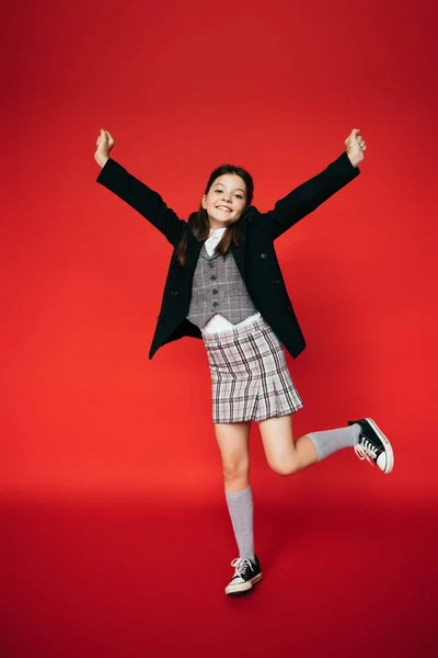 Volle Länge der aufgeregten Mädchen in kariertem Rock und schwarzem Blazer posiert auf einem Bein mit ausgestreckten Händen auf rotem Hintergrund — Stockfoto