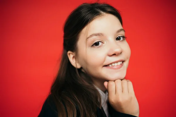 Retrato de niña preadolescente alegre sosteniendo la mano cerca de la barbilla y sonriendo a la cámara aislada en rojo - foto de stock