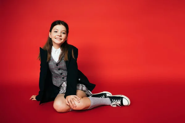 Full length of pleased girl in black blazer and gumshoes sitting on red background — Stock Photo
