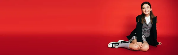 Piena lunghezza di sorridente preteen girl in giacca nera e scarpe da ginnastica seduto isolato sul rosso con spazio copia, banner — Foto stock
