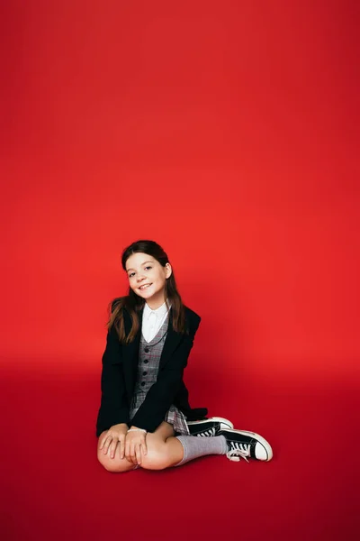Full length of stylish girl in gumshoes and black blazer smiling at camera while sitting on red background — Stock Photo