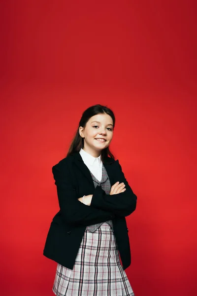 Happy brunette child in black blazer and plaid skirt standing with crossed arms isolated on red — Stock Photo