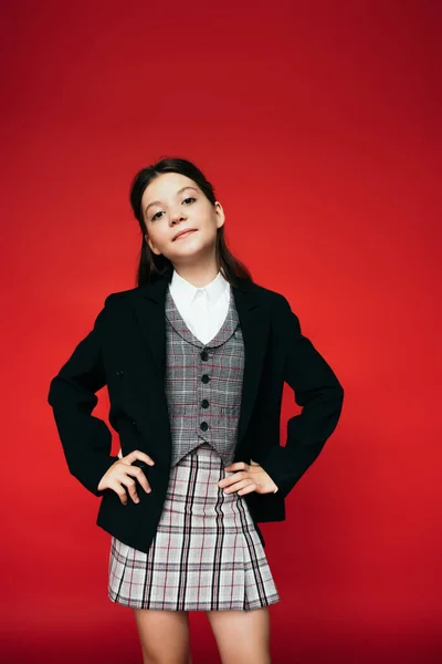 Menina positiva em colete quadriculado e blazer preto posando com as mãos sobre os quadris isolados no vermelho — Fotografia de Stock