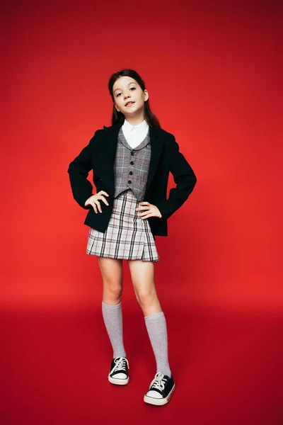 Full length of schoolgirl in black blazer and gumshoes standing akimbo on red background — Stock Photo