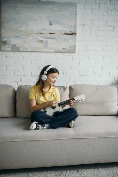 Niño positivo en auriculares sentados en un sofá moderno con las piernas cruzadas y jugando ukelele - foto de stock