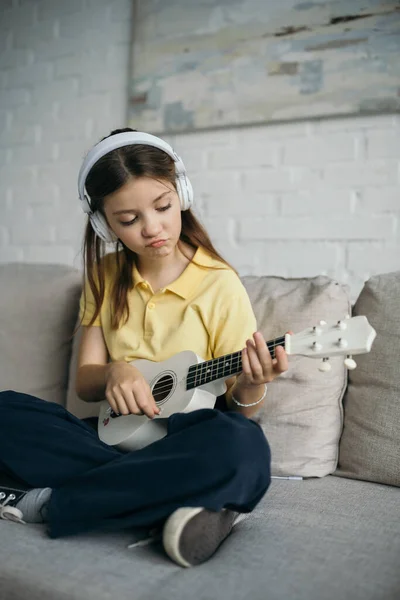 Menina descontente em fones de ouvido tocando pouca guitarra havaiana enquanto sentado no sofá com pernas cruzadas — Fotografia de Stock