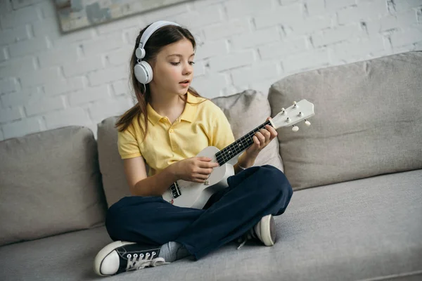 Девушка в проводных наушниках сидит на диване со скрещенными ногами и играет на укулеле — стоковое фото