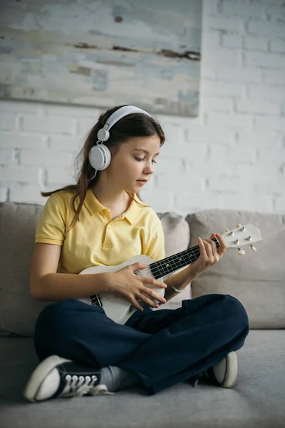 Девочка-подросток в проводных наушниках играет на укулеле, сидя со скрещенными ногами на диване — стоковое фото