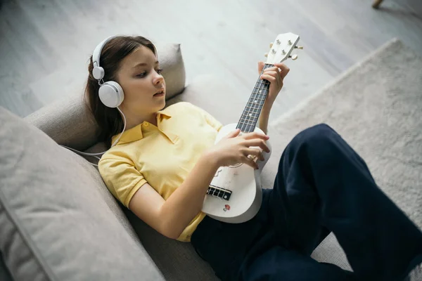 Vista superior da menina em fones de ouvido com fio deitado no sofá e tocando pequena guitarra havaiana — Fotografia de Stock