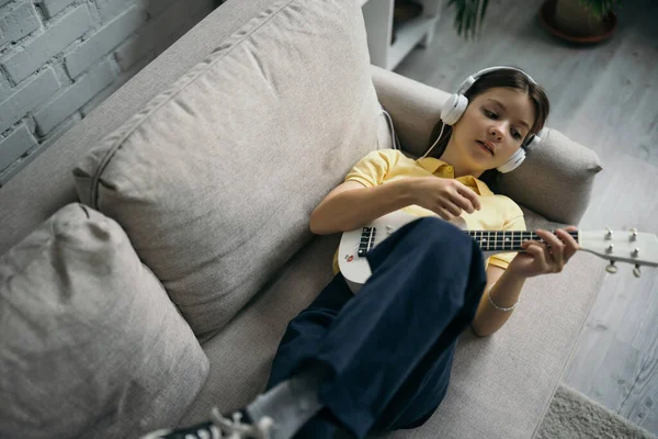 Top view of girl playing ukulele while lying on couch in wired headphones — Stock Photo
