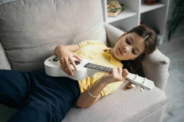 Vista de ángulo alto de la niña preadolescente acostada en el sofá en casa y tocando la pequeña guitarra hawaiana - foto de stock