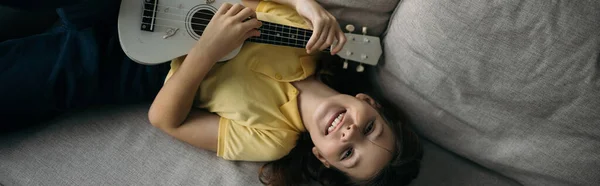 Вид зверху весела дівчина лежить на дивані і грає на маленькій гавайській гітарі, дивлячись на камеру, банер — стокове фото