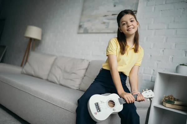 Feliz menina pré-adolescente segurando ukulele e olhando para a câmera perto do sofá embaçado em casa — Fotografia de Stock