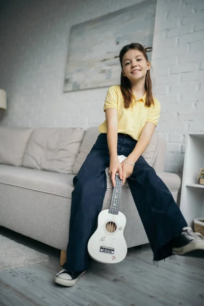 Приятная дочка с маленькой гавайской гитарой, смотрящая на камеру возле дивана в гостиной — стоковое фото