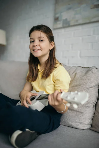 Menina pré-adolescente alegre sorrindo para a câmera enquanto joga ukulele no sofá na sala de estar — Fotografia de Stock