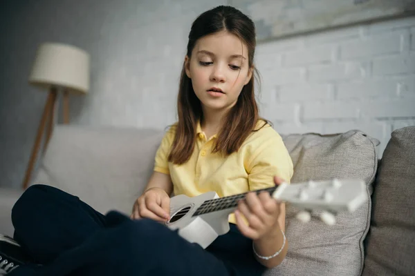 Menina morena jogando ukulele enquanto sentado no sofá em casa — Fotografia de Stock