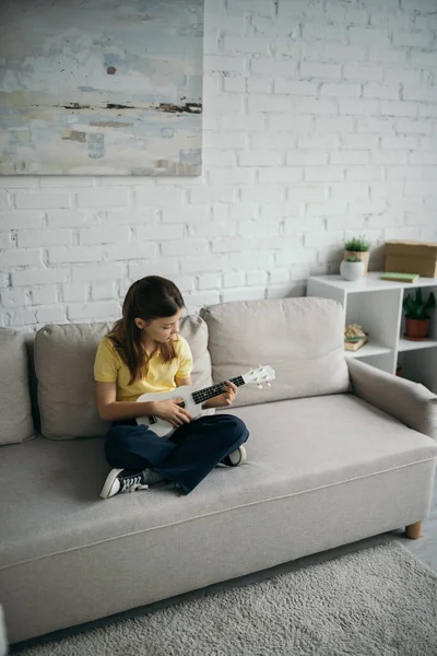 High angle view of girl sitting on modern couch with crossed legs and playing ukulele — Stock Photo