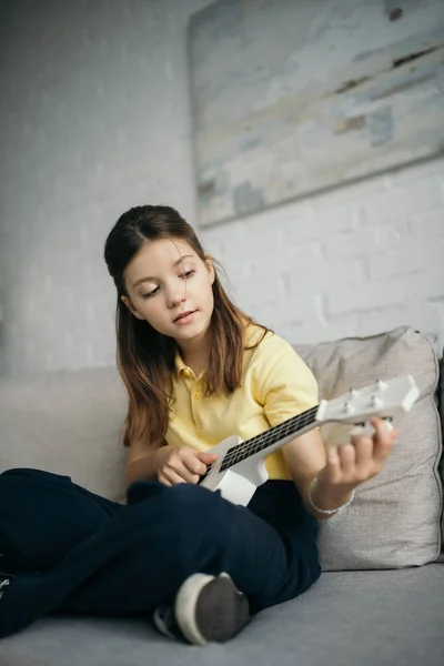 Morena menina tuning pouco havaiano guitarra enquanto sentado no sofá em casa — Fotografia de Stock