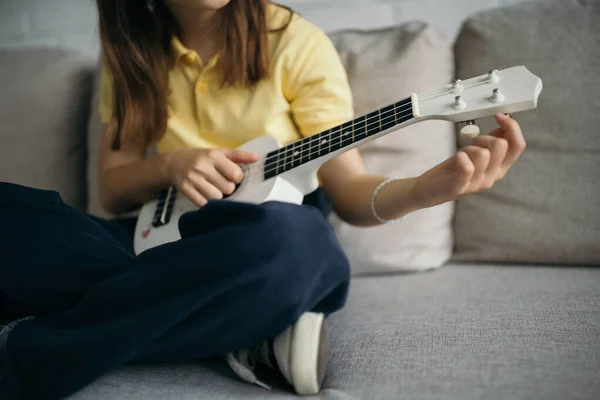 Cropped view of blurred child tuning ukulele while sitting on sofa with crossed legs — Stock Photo