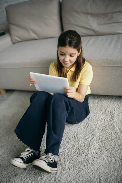 Vue grand angle de fille souriante avec tablette numérique assise sur le sol près du canapé — Photo de stock