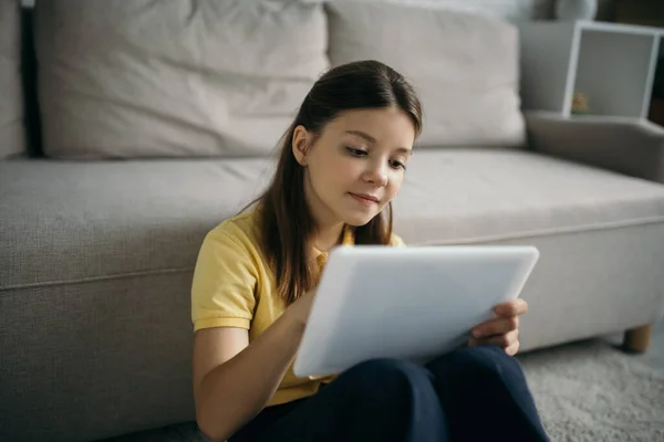 Positivo menina pré-adolescente usando tablet digital perto de sofá embaçado na sala de estar — Fotografia de Stock