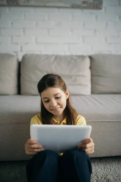 Fröhliches brünettes Mädchen blickt auf digitales Tablet in der Nähe von Couch auf verschwommenem Hintergrund — Stockfoto