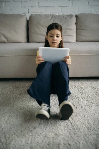 Longitud completa de la chica con la tableta digital sentado en la alfombra del piso cerca del sofá - foto de stock
