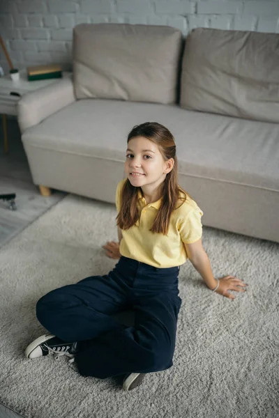 Высокий угол зрения счастливая девушка сидит на полу со скрещенными ногами возле дивана в гостиной — стоковое фото