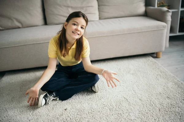 Hochwinkel-Ansicht des fröhlichen Mädchens auf dem Boden sitzend mit überkreuzten Beinen und offenen Handflächen in der Nähe von Couch zu Hause — Stockfoto