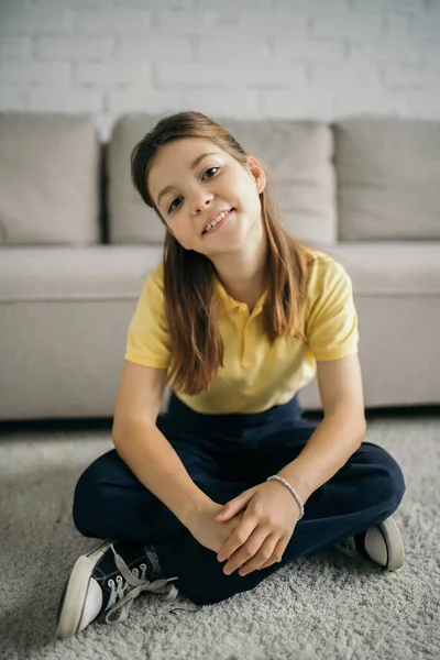 Preteen girl sitting on floor near blurred couch and smiling at camera — Stock Photo